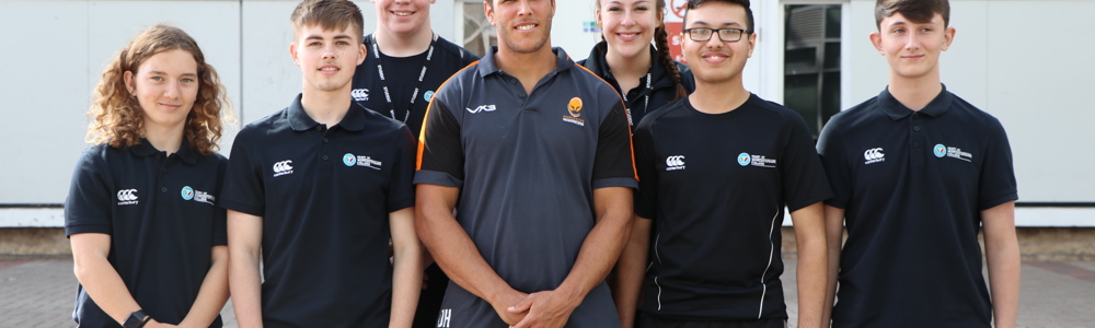 Group of 6 smiling sports students wearing matching college polo tops. Male adult smiling in the centre of the group and wearing a rugby shirt with the Worcester Warriors logo on.