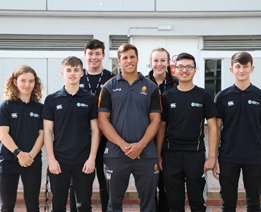 Group of 6 smiling sports students wearing matching college polo tops. Male adult smiling in the centre of the group and wearing a rugby shirt with the Worcester Warriors logo on.