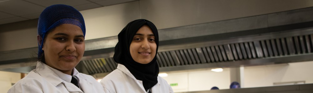 Two female students in white coats stood smiling in a commercial kitchen with food placed in front of them.