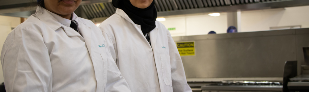 Two female students in white coats stood smiling in a commercial kitchen with food placed in front of them.