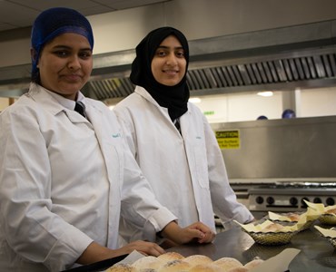 Two female students in white coats stood smiling in a commercial kitchen with food placed in front of them.