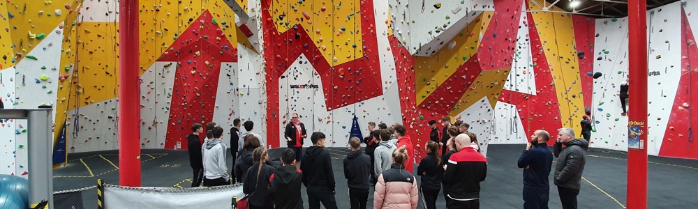 Large group of students with their back to the camera, in front of a large white, red and yellow climbing wall.