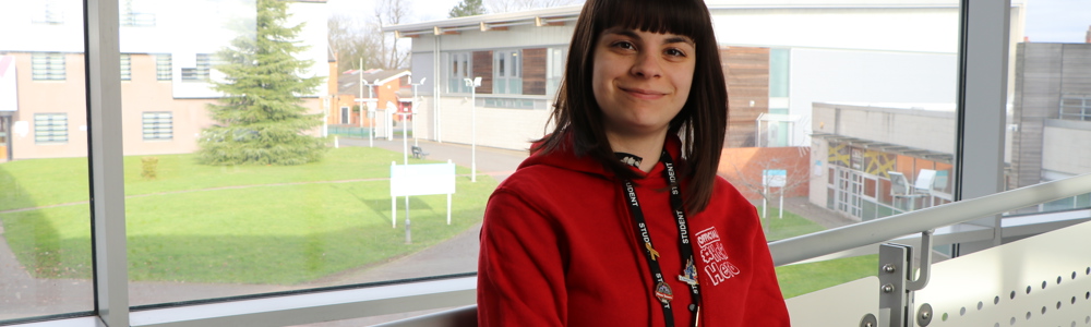 Female student in a red hoodie stood smiling in front of a large window