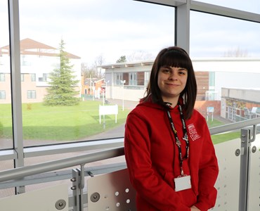 Female student in a red hoodie stood smiling in front of a large window