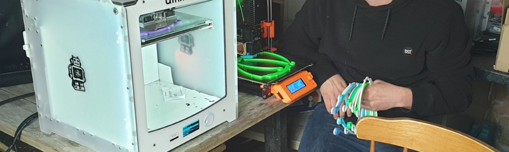 Male smiling and sat next to a 3D printer whilst packaging PPE headbands into boxes