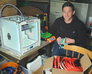 Male smiling and sat next to a 3D printer whilst packaging PPE headbands into boxes