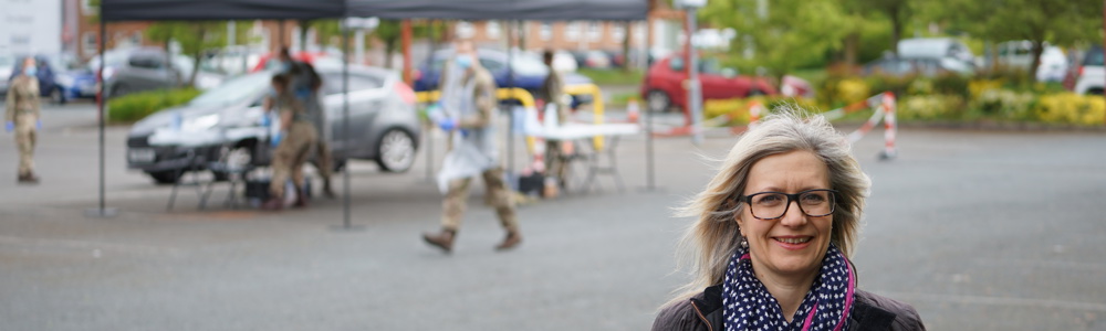 Female stood smiling in a car park with members of the army in the background directing people for Coronavirus testing