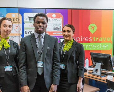 3 students in travel and tourism uniforms stood in Heart of Worcestershire College's Spires Travel Agency smiling at the camera