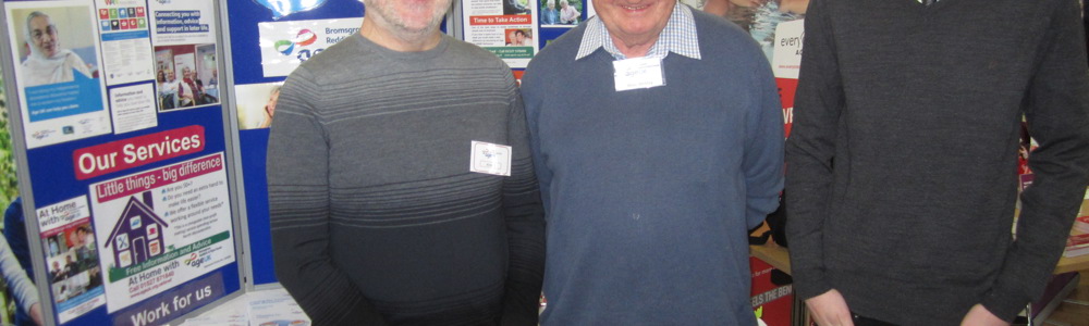 1 male student and 2 senior males stood in front of a stall at a fair.