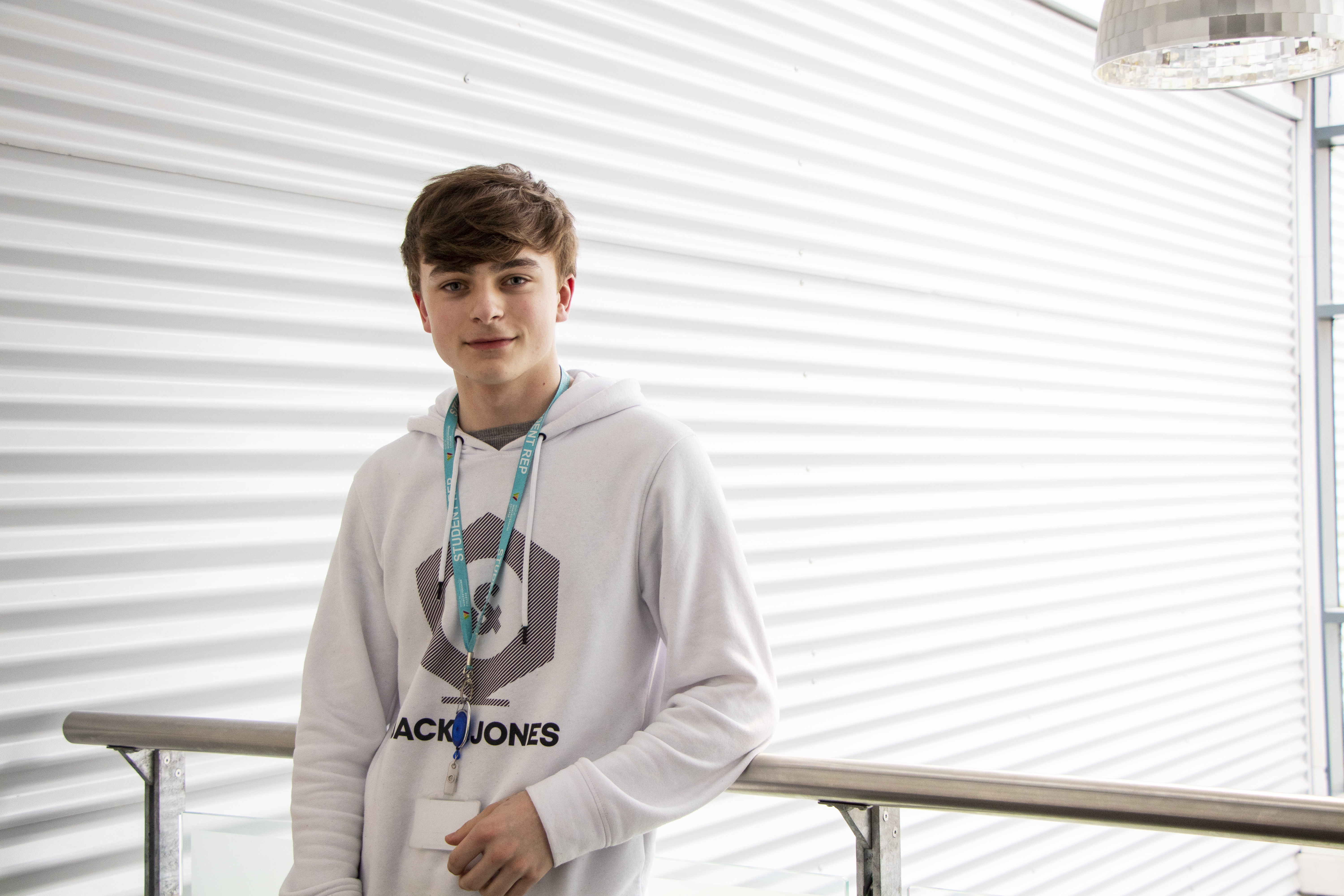 Male student in a white hoodie stood smiling in front of a indoor balcony rail