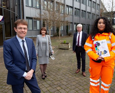 4 adults stood socially distanced outside the front of a College. 2 males wearing suits, one female wearing business attire and one female wearing orange high visibility jacket and trousers while holding a 'reignite your future' brochure.