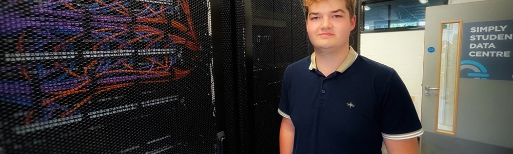 Male students stood against a computer server wall smiling at the camera