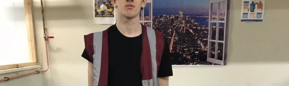 Image of male plumbing student wearing a high visibility jacket, hard hat, and stood in front of some pipes and a sink.