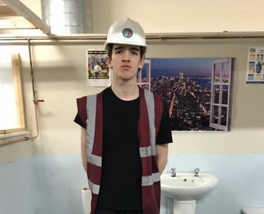 Image of male plumbing student wearing a high visibility jacket, hard hat, and stood in front of some pipes and a sink.
