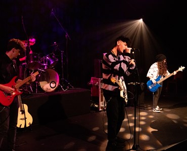 A group of students on a stage performing; two students are playing guitars, one is singing into microphones and one student is playing the drums