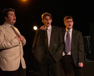 A performing arts dress rehearsal. 3 males students dressed in suits, singing on stage.