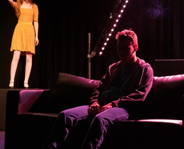 A performing arts dress rehearsal. A male student sat on a sofa with a female student in a yellow dress stood in the background
