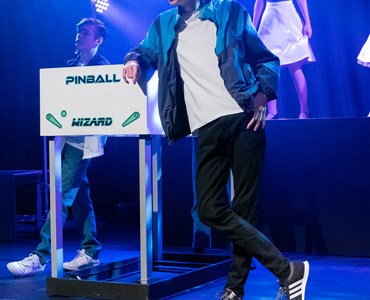 A performing arts dress rehearsal. 2 male students dressed in jeans, trainers and varsity jacket, leaning against a post. A female student is stood on a podium behind