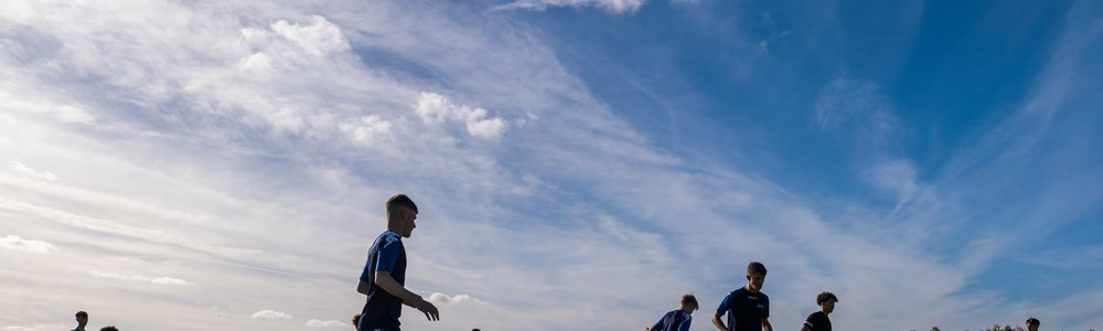 Male students undertake football training on an outdoor grass pitch under blue skies and sunshine