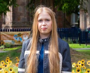 Front facing shot of T Level student Courtney who stares directly into the camera, her long blonde hair drapes over her shoulders and sunflowers can be seen in the background on a bright summers day