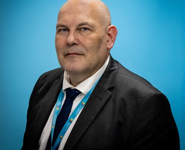 picture of Simon Kibble in front of a blue background wearing a suit and tie